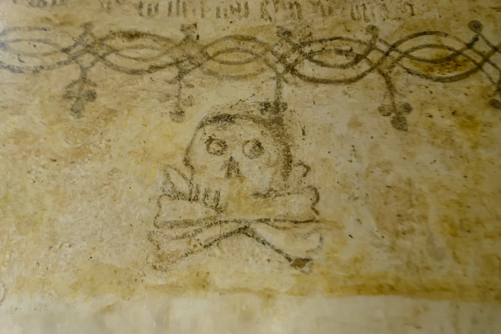 A skull and crossbones painted in a bold, simple style on the lime-washed east wall near the altar.